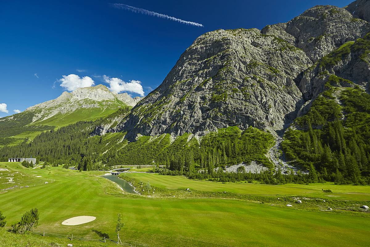 Ein weiterer sonniger Ausblick auf den smaragdgrünen Golfclub Lech, zusammen mit Bergen als Hintergrund-Kulisse.