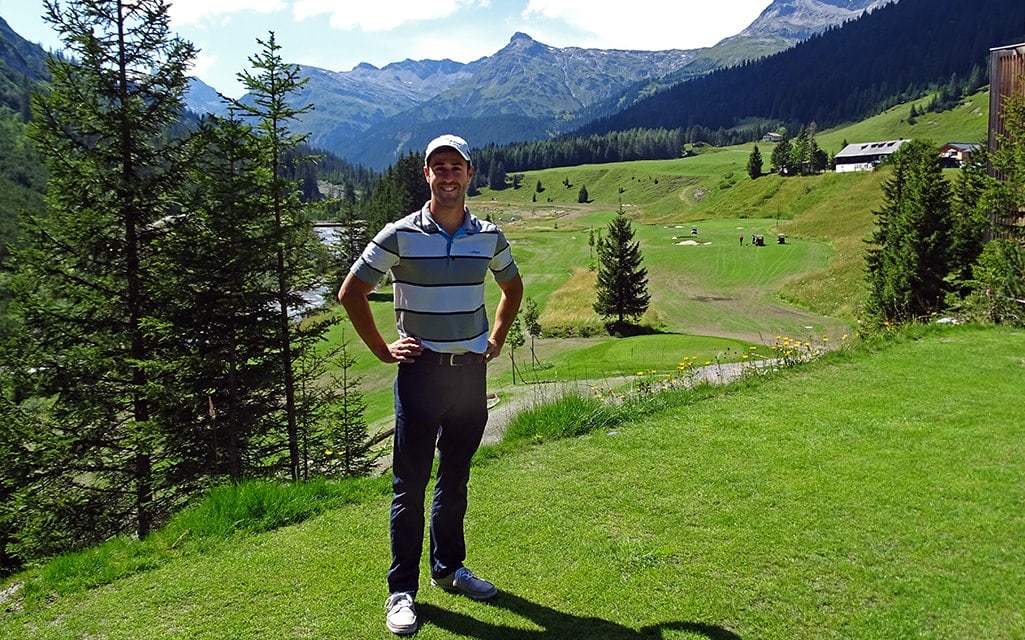 Mann & Golf-Pro in Golfbekleidung steht Modell beim smaragdgrünen Golfplatz mit Bergen im Hintergrund.