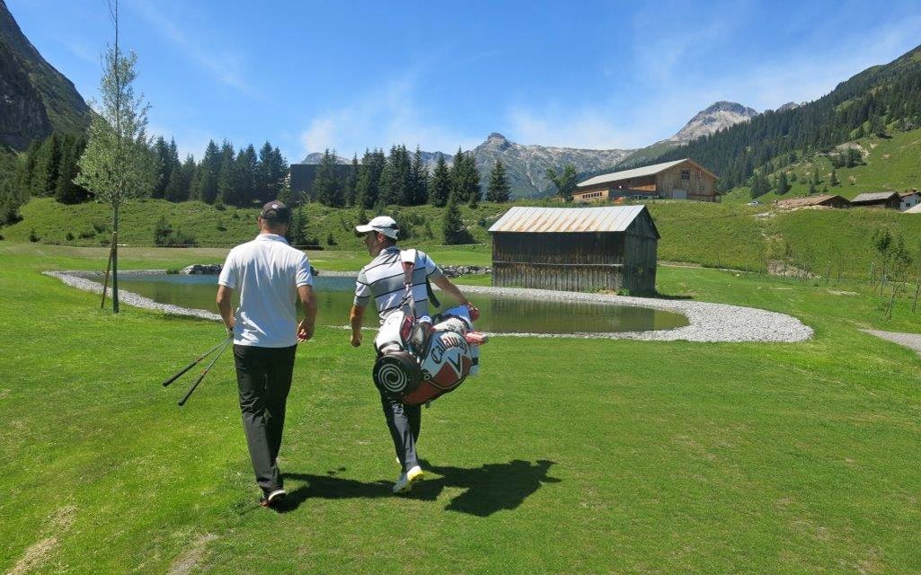 Two men carrying golf clubs on the course with the mountains in the background.