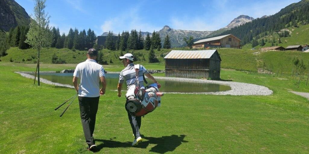 Two men carrying golf clubs on the course with the mountains in the background.