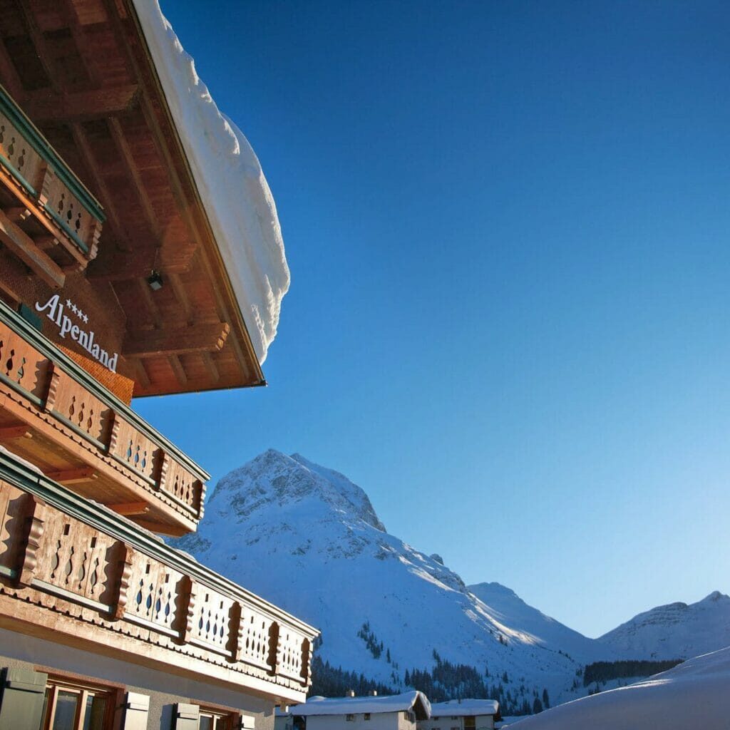 Teilweise Aussenansicht und Perspektive vom Hotel Alpenland mit einem schneebedeckten Platz an einem sonnigen Tag.